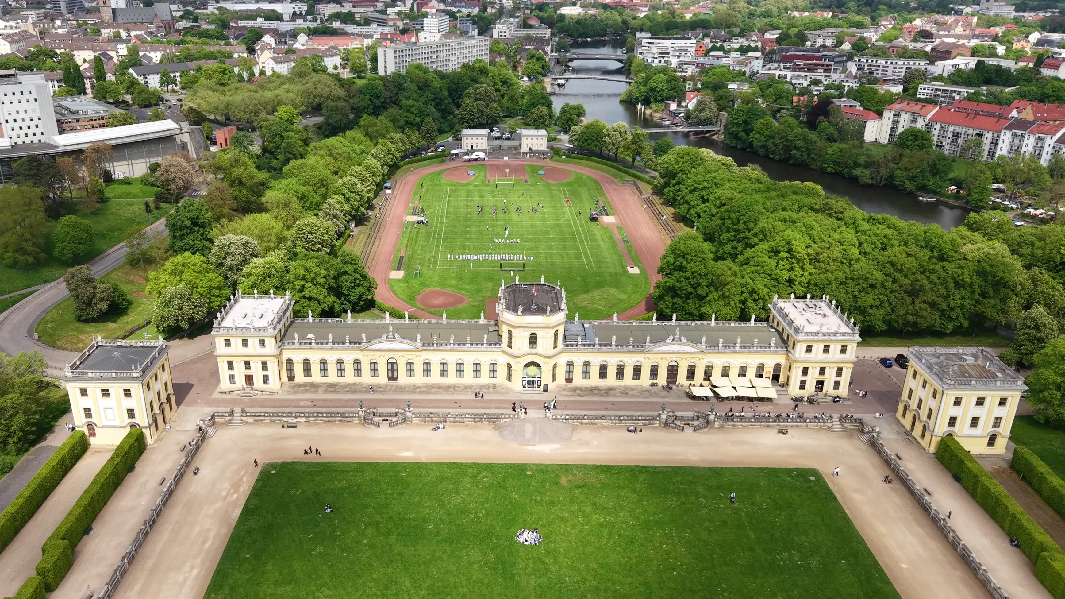 Langes barockes Haus mit zwei Seitenpavillons aus der Luft, davor ein Rasenparterre, dahinter Park, Fluss und Stadt