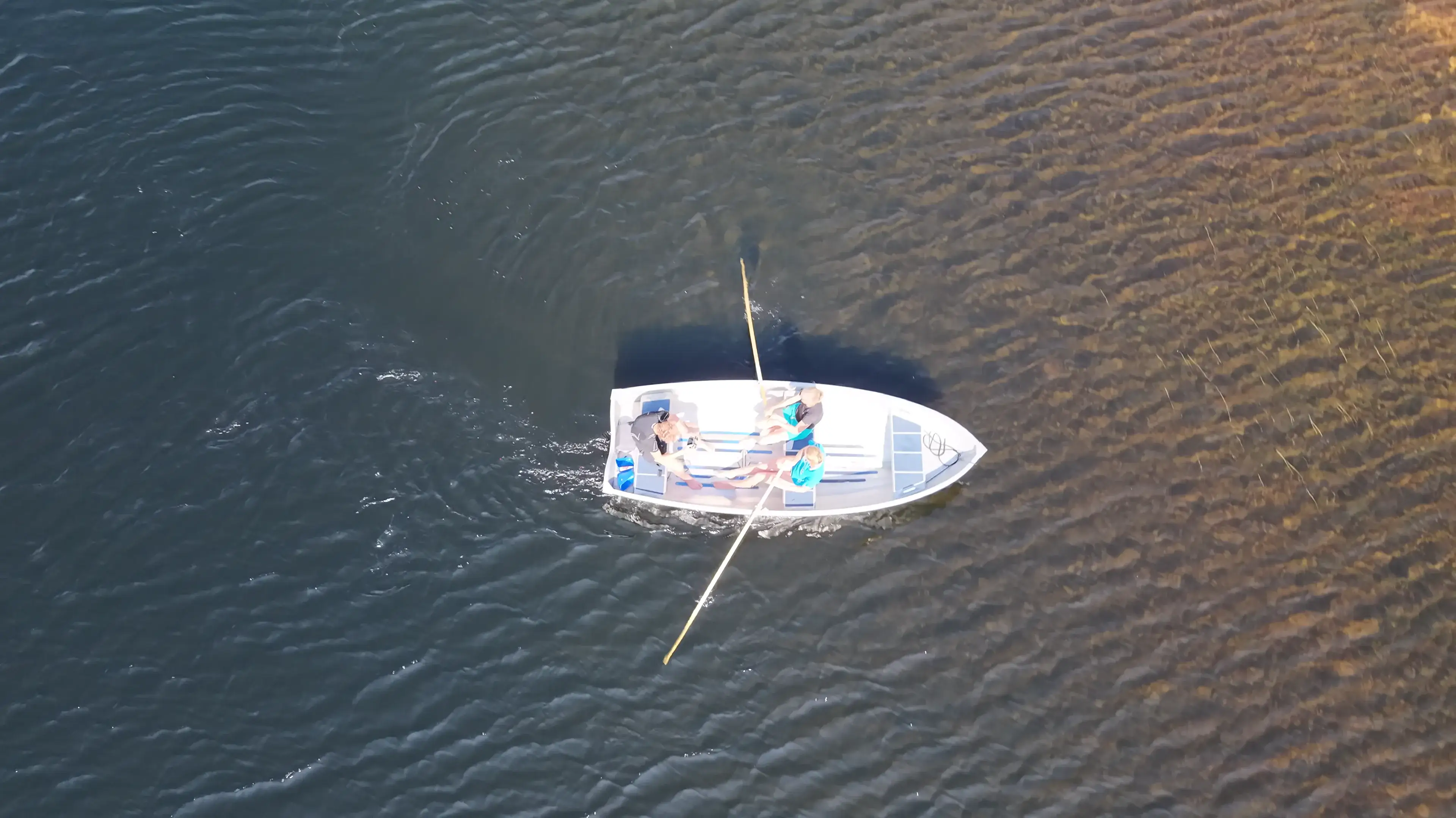 Ein kleines Ruderboot mit drei Personen, direkt von oben über flachem, klarem Wasser. Die Kamera weicht zurück und gibt das steinige, moosgrüne Ufer dahinter frei.
