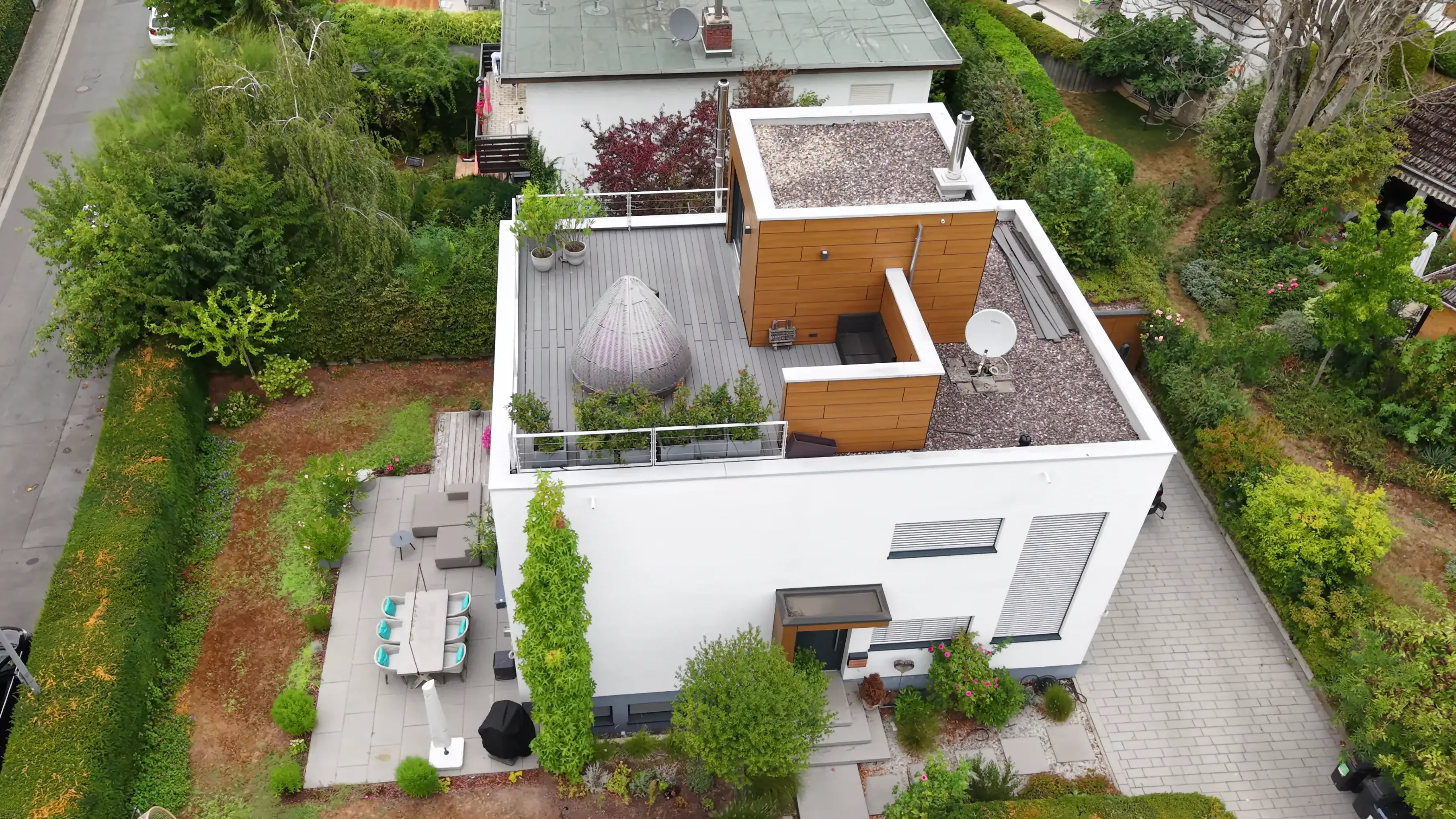 Schräge Luftaufnahme eines weißen Flachdachhauses: Dachterrasse mit Lounge-Sessel und Pflanzkübeln, darunter die Gartenterrasse mit Esstisch, dahinter Hecken, Nachbargärten und die gepflasterte Zufahrt.